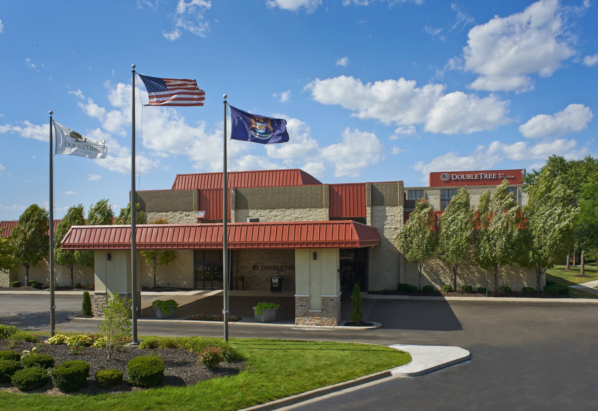 The porte cochere of the Detroit-Dearborn DoubleTree by Hilton Hotel. In front of the entrance are three flagpoles displaying three different flags: the DoubleTree by Hilton flag, the flag of the United States, and the state flag of Michigan.